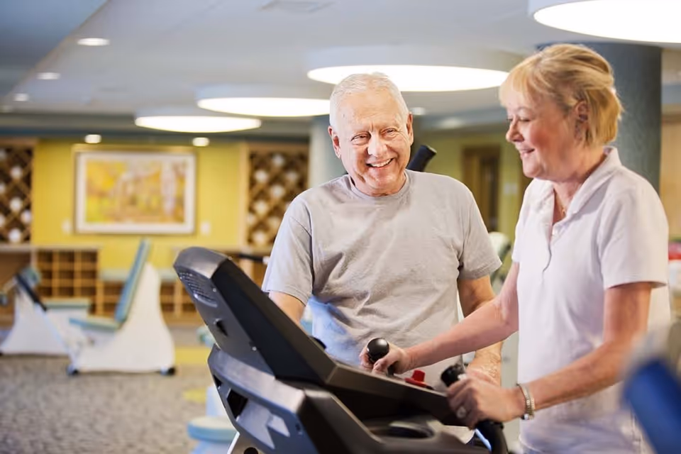 An elderly man and a woman smiling and interacting while using exercise equipment in a fitness area of a senior living community.