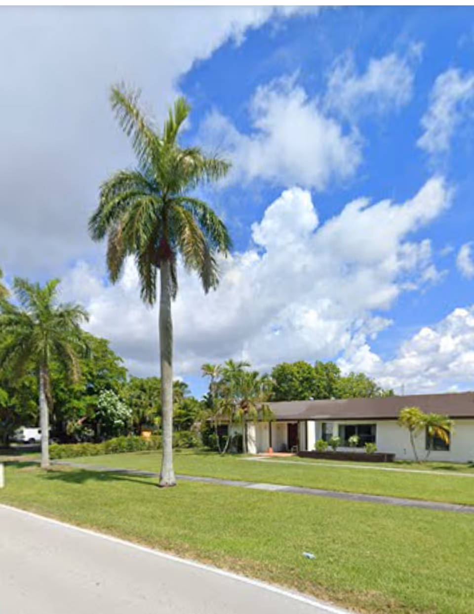 Front exterior of a single-story retirement home with palm trees, a grassy lawn, and a bright cloudy sky.