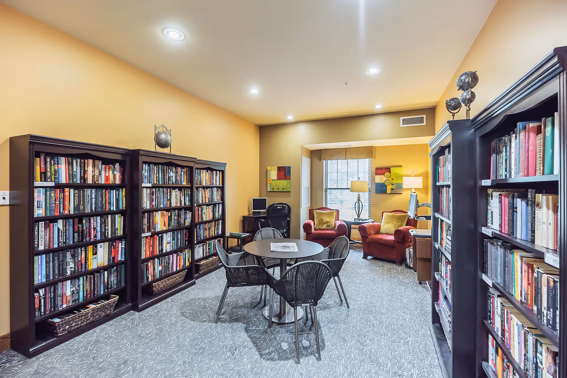 Cozy senior living library/lounge with bookshelves, a round table and chairs, and two upholstered armchairs by a window.