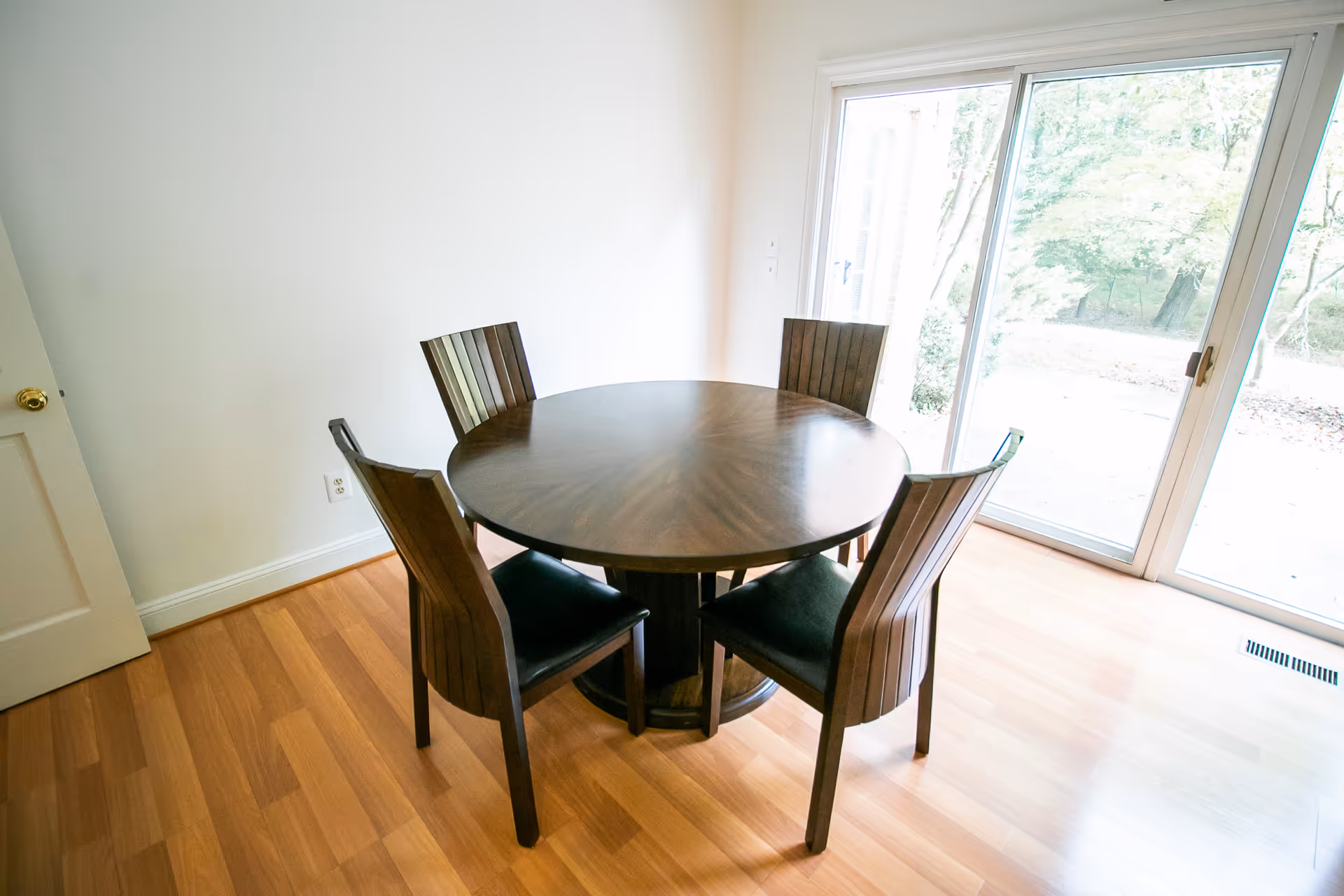 A bright dining area with a round wooden table surrounded by four matching wooden chairs with black cushions. The room has light-colored walls and wood flooring, with a large sliding glass door letting in natural light and showing an outdoor view.