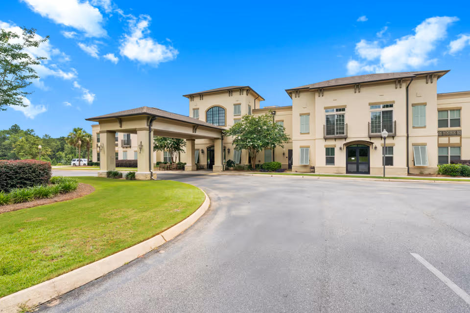 Exterior view of Seabrook Pointe facility showing a beige two-story building with large windows and a covered entrance driveway. The surrounding area includes a well-maintained lawn, shrubs, trees, and a clear blue sky with some clouds.