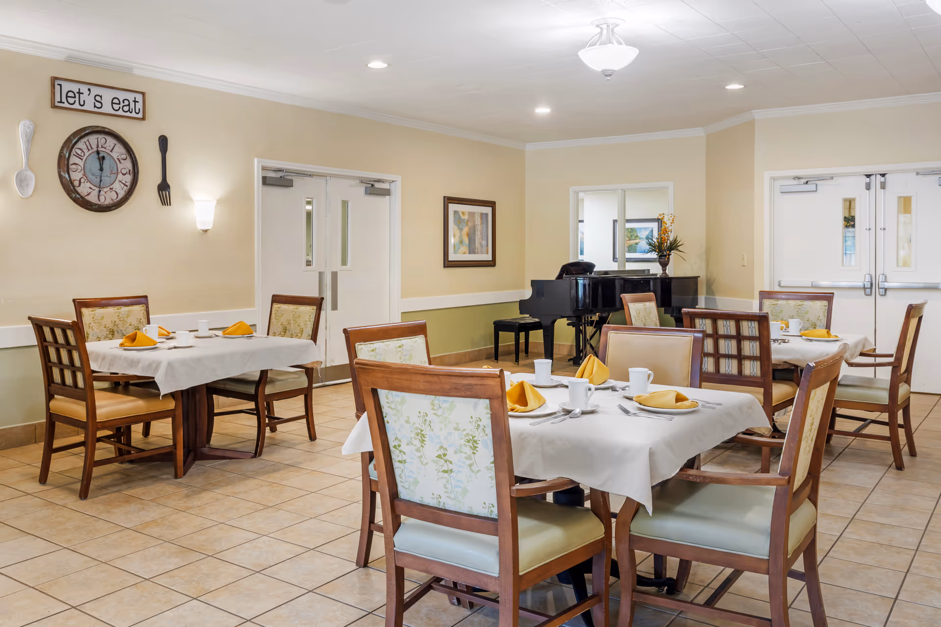 A dining room with several tables covered with white tablecloths, each set with white cups, plates, silverware, and yellow folded napkins. The room has tiled floors, beige walls, and a black grand piano in the corner. On the wall, there is a clock and a sign that reads 'let's eat' with decorative oversized spoon and fork.