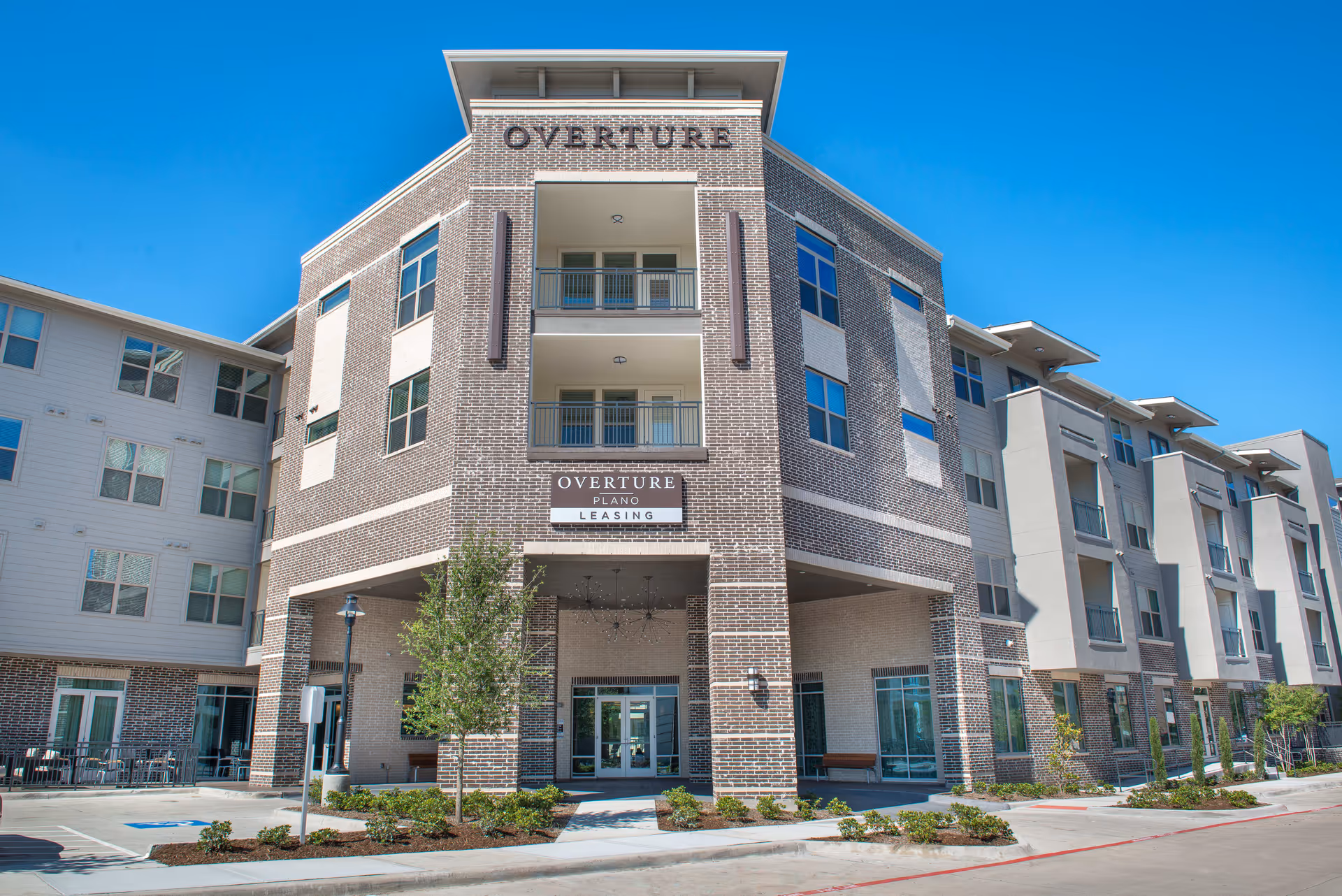 Exterior view of a modern multi-story brick and siding building with balconies and large windows under a clear blue sky. The building has a sign that reads 'OVERTURE PLANO LEASING' above the entrance, with landscaping and a parking area in front.
