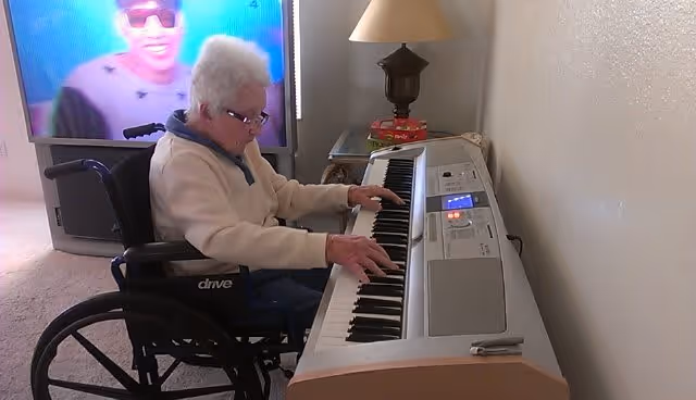 An elderly person in a wheelchair playing an electronic keyboard in a room. A television is on in the background showing a person wearing sunglasses. There is a table with a lamp and some books next to the keyboard.