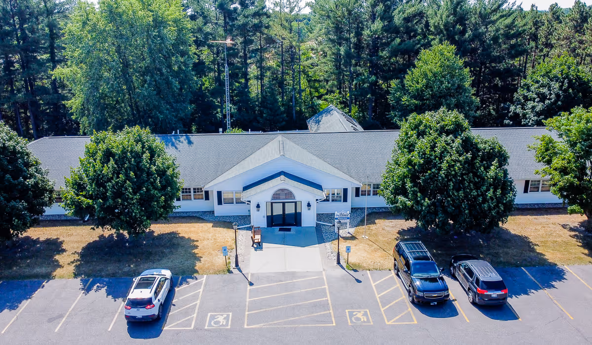 Front exterior view of a single-story senior living facility building with a gray roof and white walls, surrounded by trees. There is a paved parking lot in front with three vehicles parked, including two in accessible parking spaces. The entrance has a small covered porch with double doors.