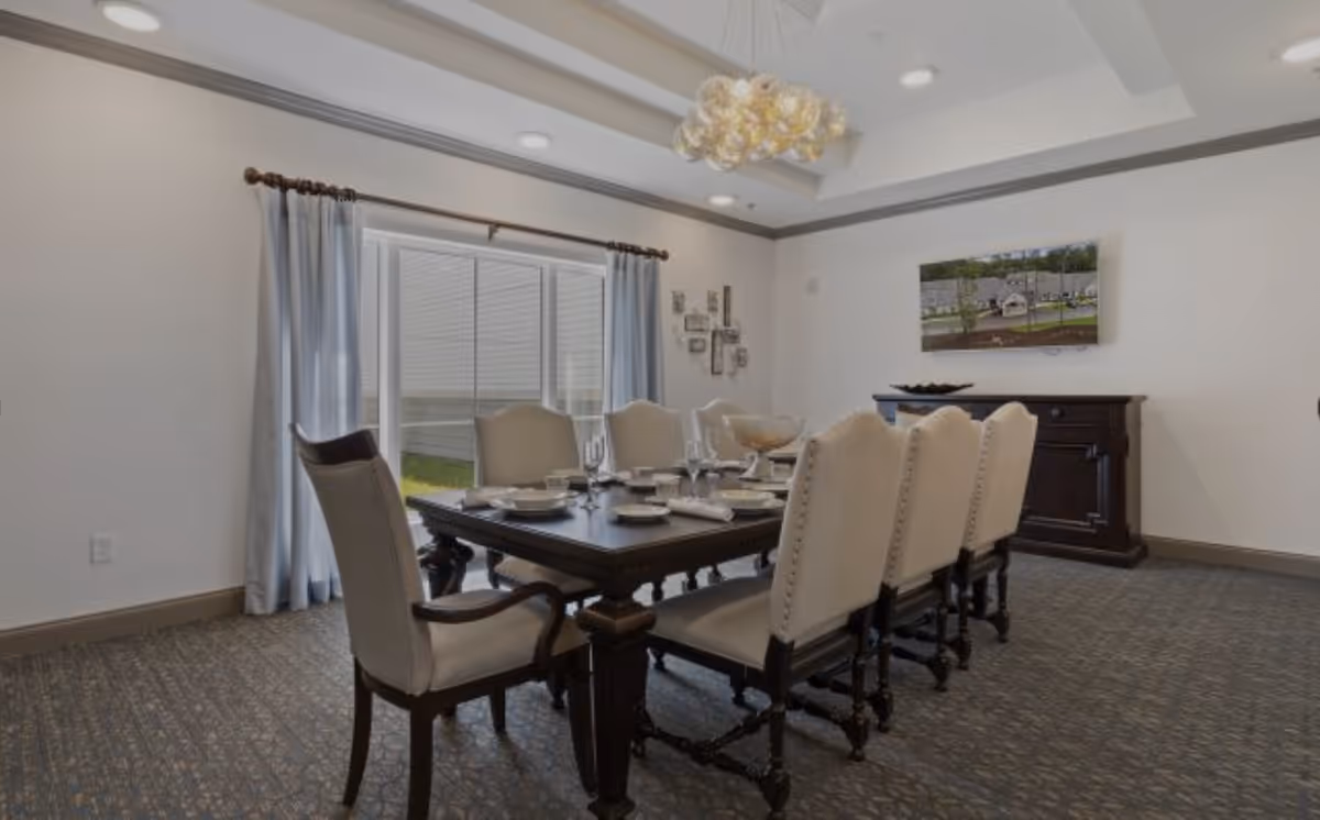 A formal dining room with a dark wooden dining table set for eight people with beige upholstered chairs. The room has light-colored walls, a large window with light blue curtains, a decorative chandelier, and a sideboard with a framed picture above it.