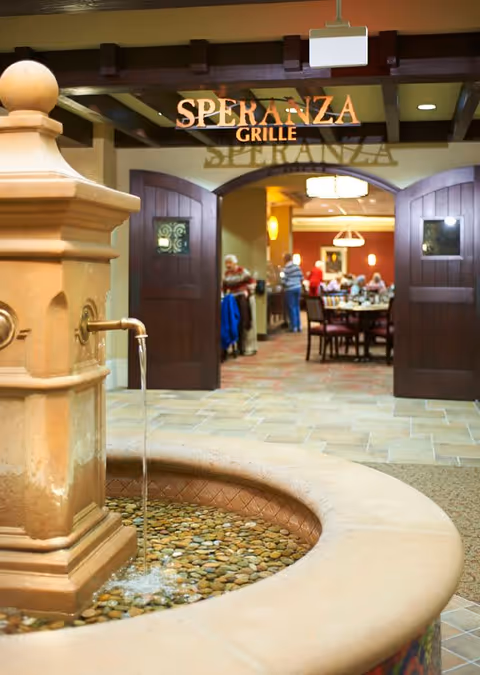 Indoor view of a water fountain with flowing water in the foreground, leading to an open double door entrance to a dining area named Speranza Grille, where several people are seated and standing.
