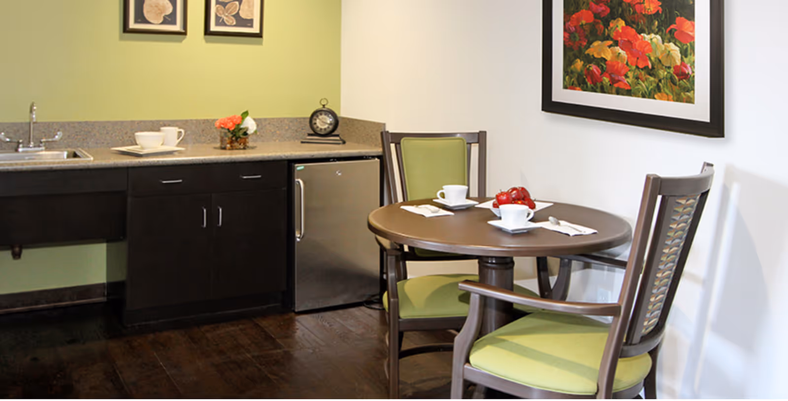 A small dining area with a round wooden table set for two with white cups and napkins. Two wooden chairs with green cushions surround the table. To the left, there is a kitchenette with a sink, dark cabinets, a small refrigerator, and a countertop decorated with a small flower arrangement and a clock. The walls are painted light green and white, with framed artwork hanging above the table and countertop.