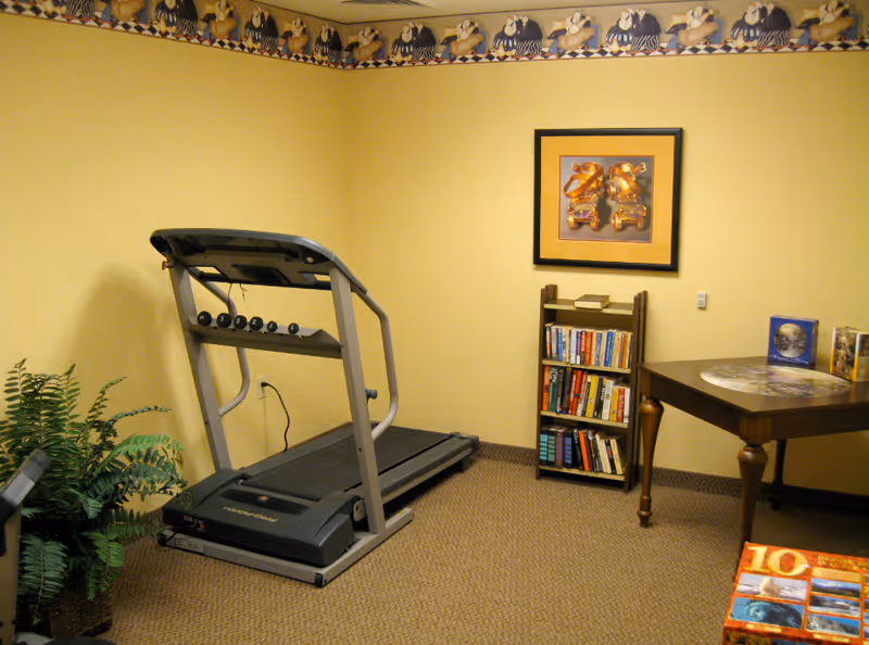 A small room with a treadmill, a bookshelf filled with books, a wooden table with books and a decorative placemat, a potted plant, and a framed picture on the wall. The walls are painted yellow with a decorative border near the ceiling featuring ducks.