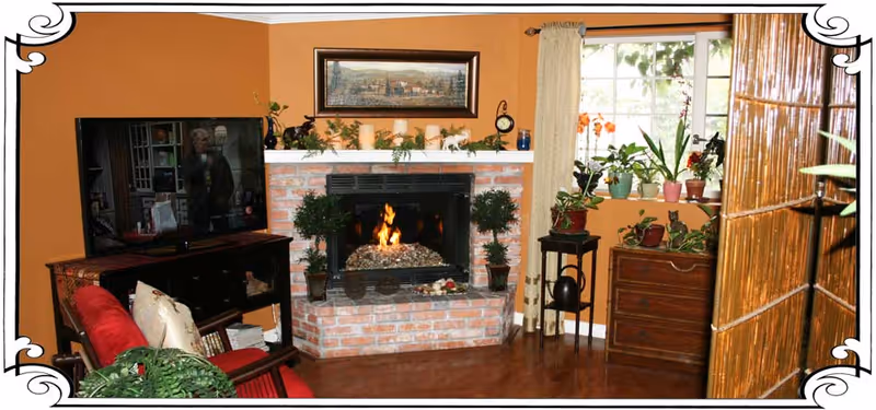Cozy living room with a brick fireplace lit with a fire, flanked by two potted plants. Above the fireplace is a framed landscape painting and decorative candles. To the left, there is a flat-screen TV on a dark wooden stand, and a red cushioned chair with pillows. To the right, a window with cream curtains lets in natural light, and several potted plants are placed on a wooden chest and a small stand. A bamboo room divider is partially visible on the right side of the image.