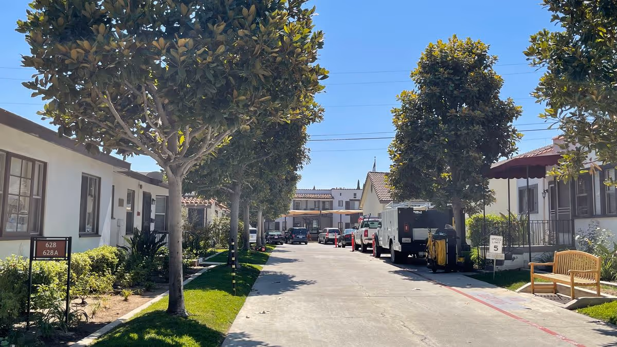 Tree-lined driveway between single-story residential buildings with parked vehicles and a bench.