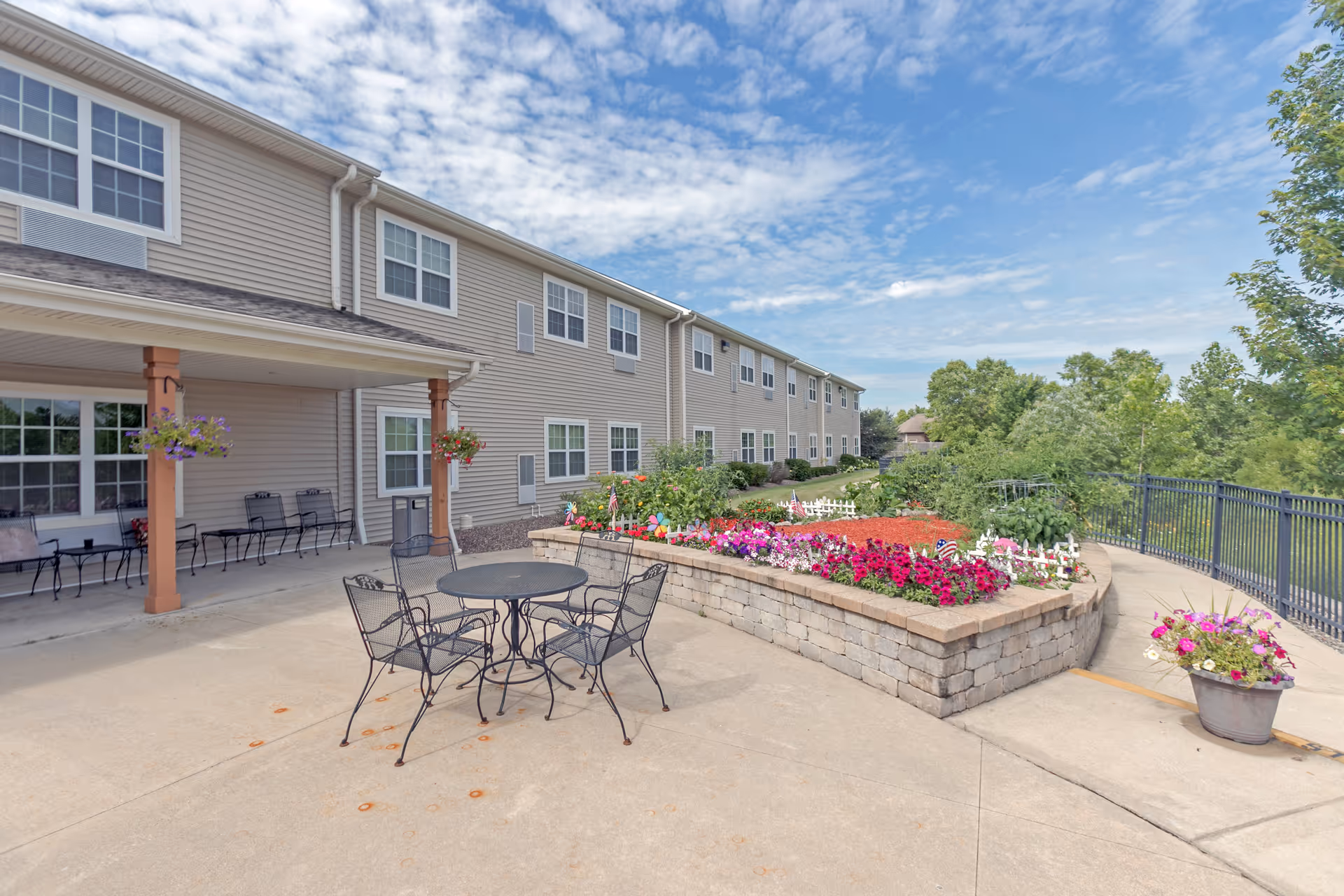 Outdoor patio area at Heritage Woods of Yorkville featuring a round metal table with four chairs, a raised flower bed with colorful flowers, hanging flower pots, and a two-story building with multiple windows in the background under a partly cloudy sky.