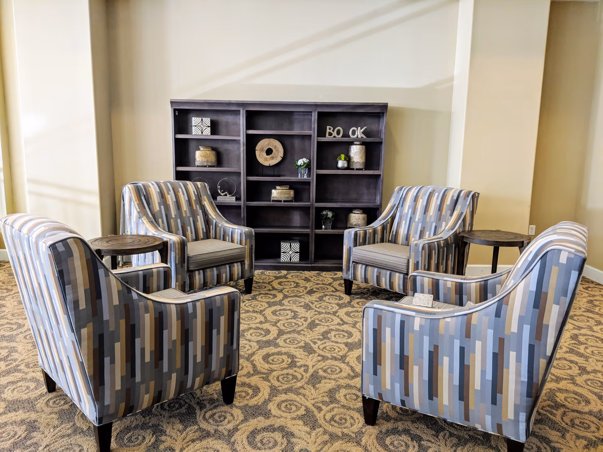 A seating area with four patterned armchairs arranged in a circle around two small round wooden side tables, set on a patterned carpet. Behind the chairs is a dark wooden bookshelf with decorative items and letters spelling 'BOOK'. The walls are painted beige.