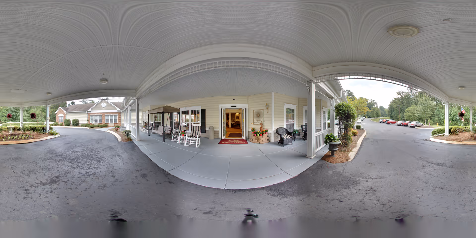 Covered porte-cochere entrance of a senior living facility with rocking chairs, a swing, landscaped beds and a driveway with parked cars.