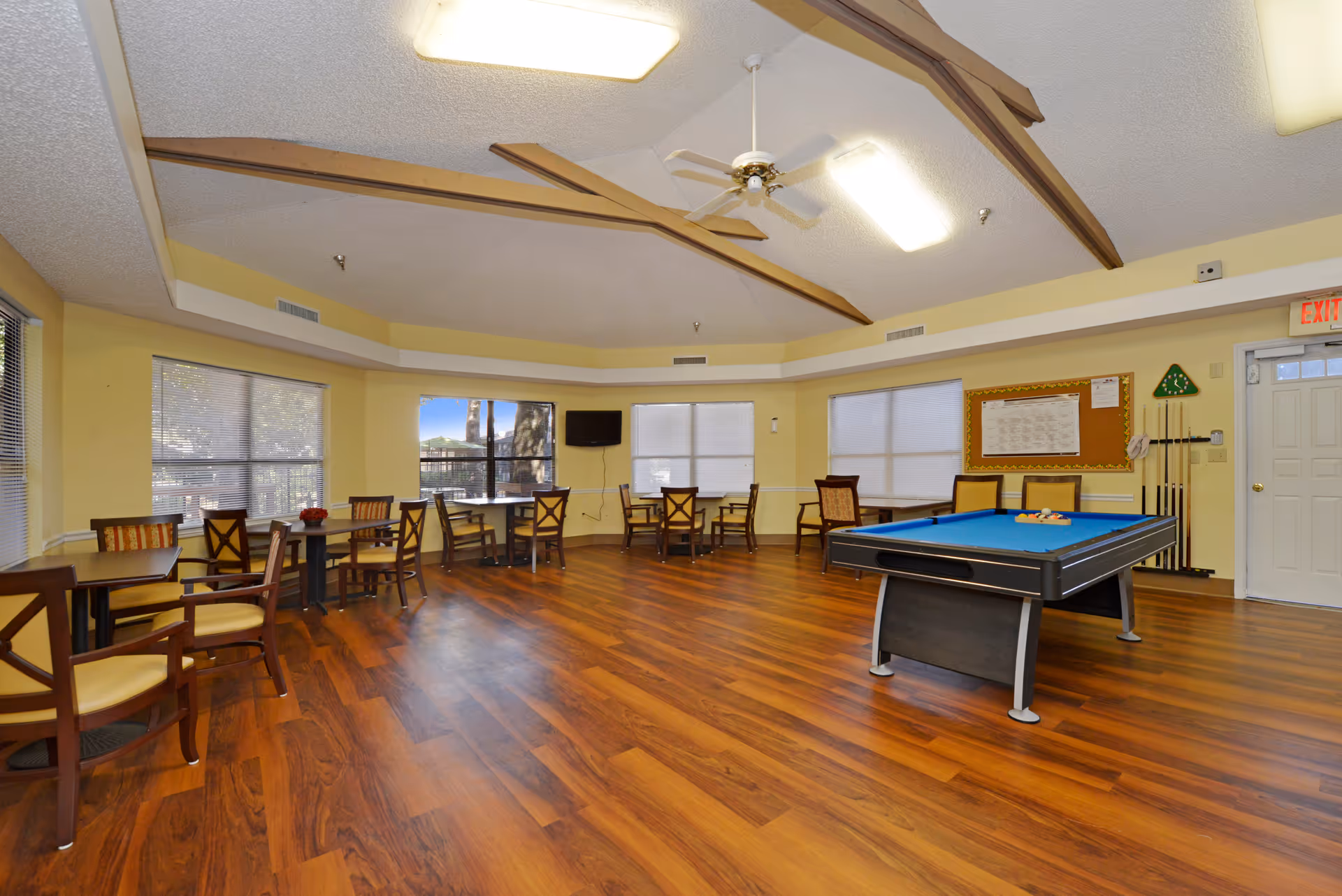 A spacious common room with wooden flooring, several tables and chairs arranged around the room, a pool table with billiard balls set up, large windows with blinds, a wall-mounted TV, and a bulletin board. The ceiling has wooden beams and a ceiling fan, and there is an exit door on the right side.