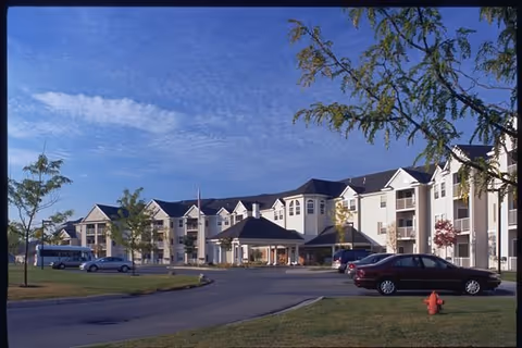 Exterior view of a large, multi-story senior living facility named Grandhaven Manor with a covered entrance, several parked cars, and trees on a clear day with blue sky and scattered clouds.