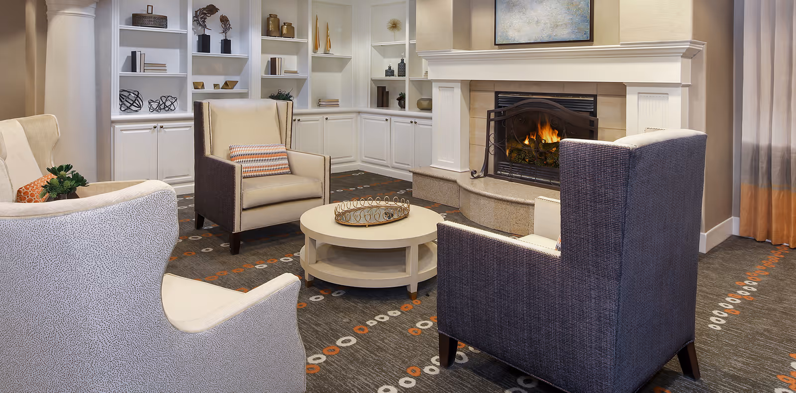 A cozy living room area with a lit fireplace, surrounded by four upholstered armchairs arranged around a round coffee table. The room features built-in white shelves with decorative items and books, a patterned carpet, and a painting above the fireplace.