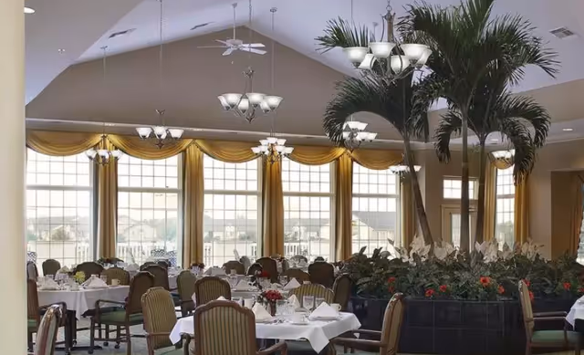Elegant dining room with tables set in white linens, chandeliers, large arched windows and indoor palm trees.