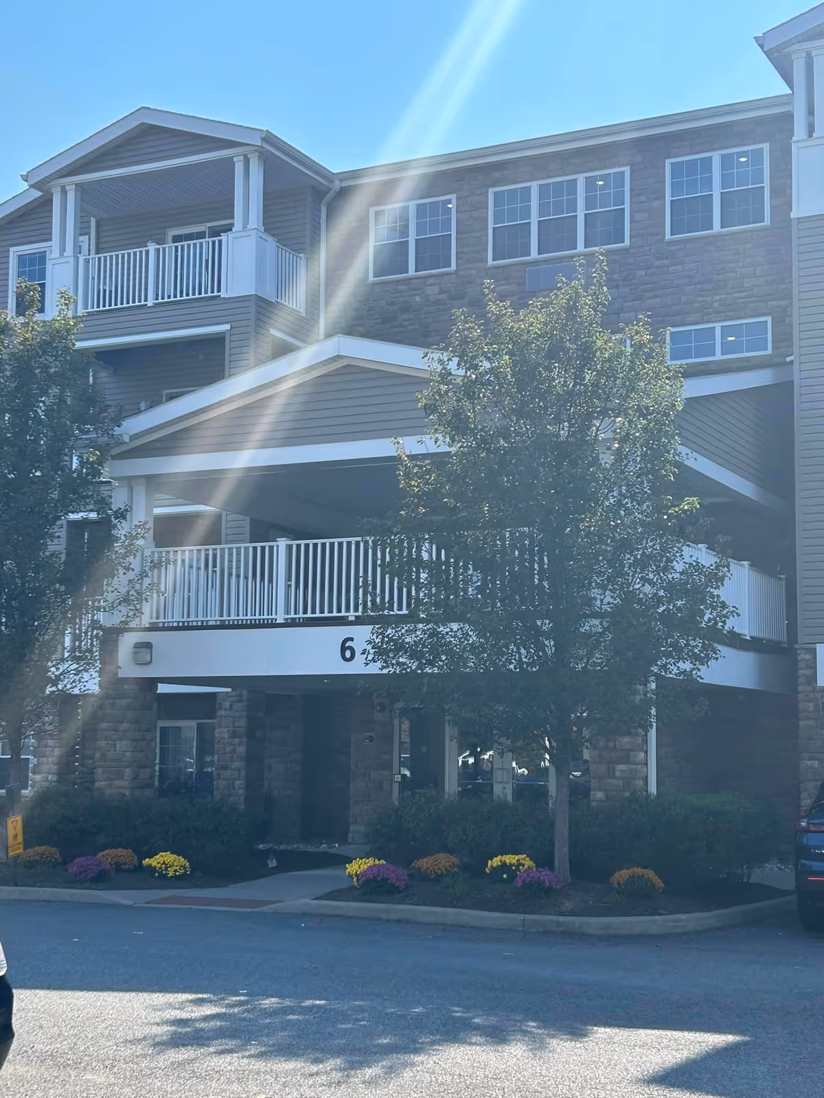 Exterior view of a multi-story senior living facility building with stone and siding facade, white railings on balconies, a covered entrance with the number 6 displayed, surrounded by trees and flower beds under a clear blue sky.