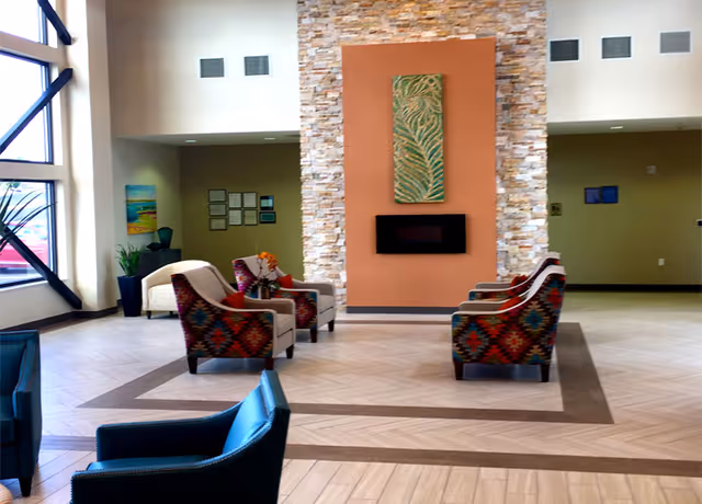 A spacious and well-lit common area with large windows on the left side. The room features a central stone accent wall with a mounted modern electric fireplace and a decorative artwork above it. Surrounding the fireplace are several upholstered armchairs with colorful patterned fabric and solid colors arranged for conversation. The floor has a light wood pattern with darker wood inlays creating a geometric design. There are plants and framed pictures on the walls in the background.