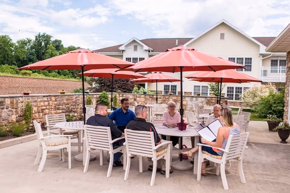 A group of six people sitting around a round outdoor table with white chairs and red umbrellas on a patio area. The background shows a stone wall, greenery, and a multi-story building with white siding and multiple windows. The sky is partly cloudy.