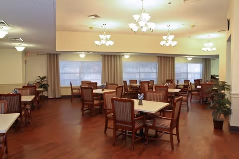 A spacious dining room with multiple square tables and wooden chairs arranged neatly. The room has large windows with beige curtains, warm lighting from ceiling chandeliers, and potted plants placed around the perimeter. The floor is wooden and the overall ambiance is clean and inviting.