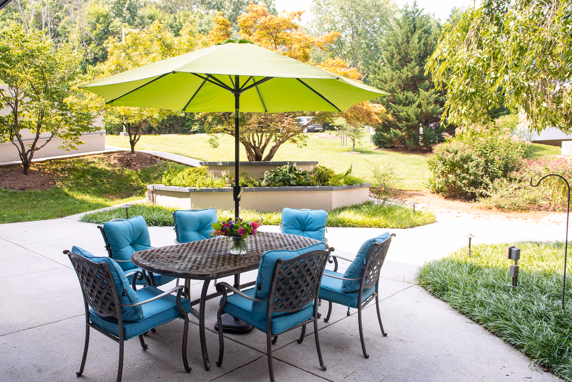 Outdoor patio with a round metal table, six blue-cushioned chairs and a large green umbrella overlooking landscaped gardens.