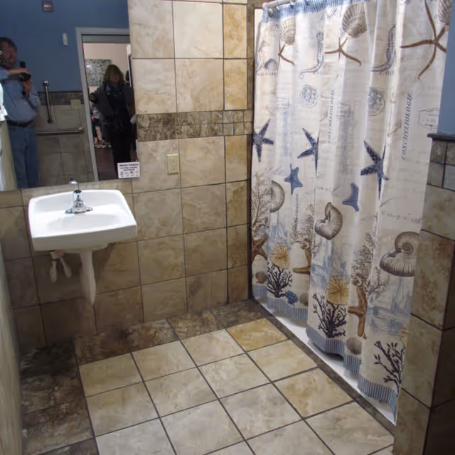 A bathroom with beige and brown tiled walls and floor, a white wall-mounted sink with a faucet, and a shower area with a curtain featuring starfish, seashells, and coral designs. A mirror on the wall reflects two people standing in the doorway.