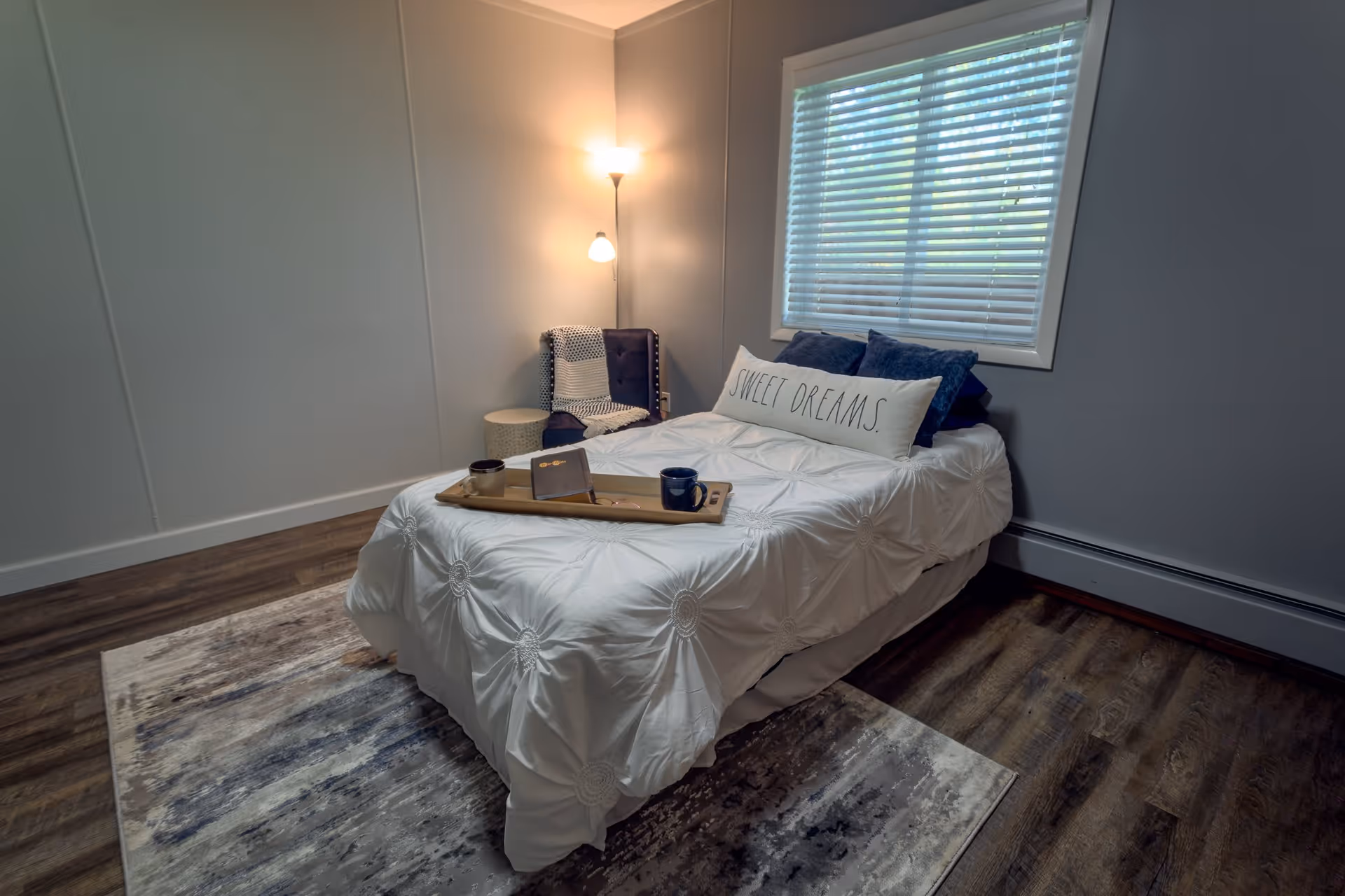 A cozy bedroom with a single bed covered in a white textured comforter and a long pillow that says 'SWEET DREAMS.' There is a wooden tray on the bed with two mugs and a book. Next to the bed is a small round table and a cushioned chair with a blanket draped over it. A floor lamp with two lights is turned on in the corner, and a window with white blinds lets in natural light. The room has wooden flooring and a patterned area rug.