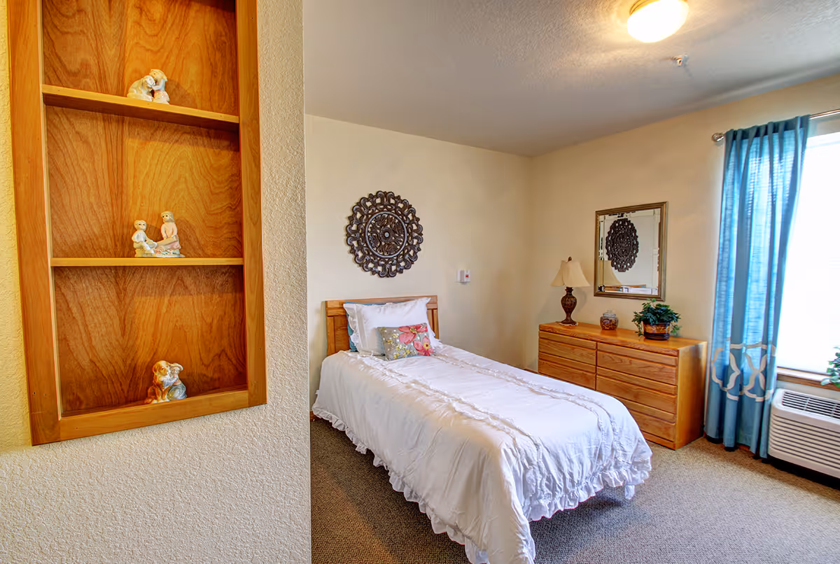 A cozy bedroom in a senior living facility featuring a single bed with white bedding and a floral pillow. There is a wooden dresser with a lamp, a plant, and a decorative jar on top. A mirror hangs above the dresser, reflecting a decorative wall piece. Blue curtains frame a window next to an air conditioning unit. On the left side, a built-in wooden shelf holds small figurines.
