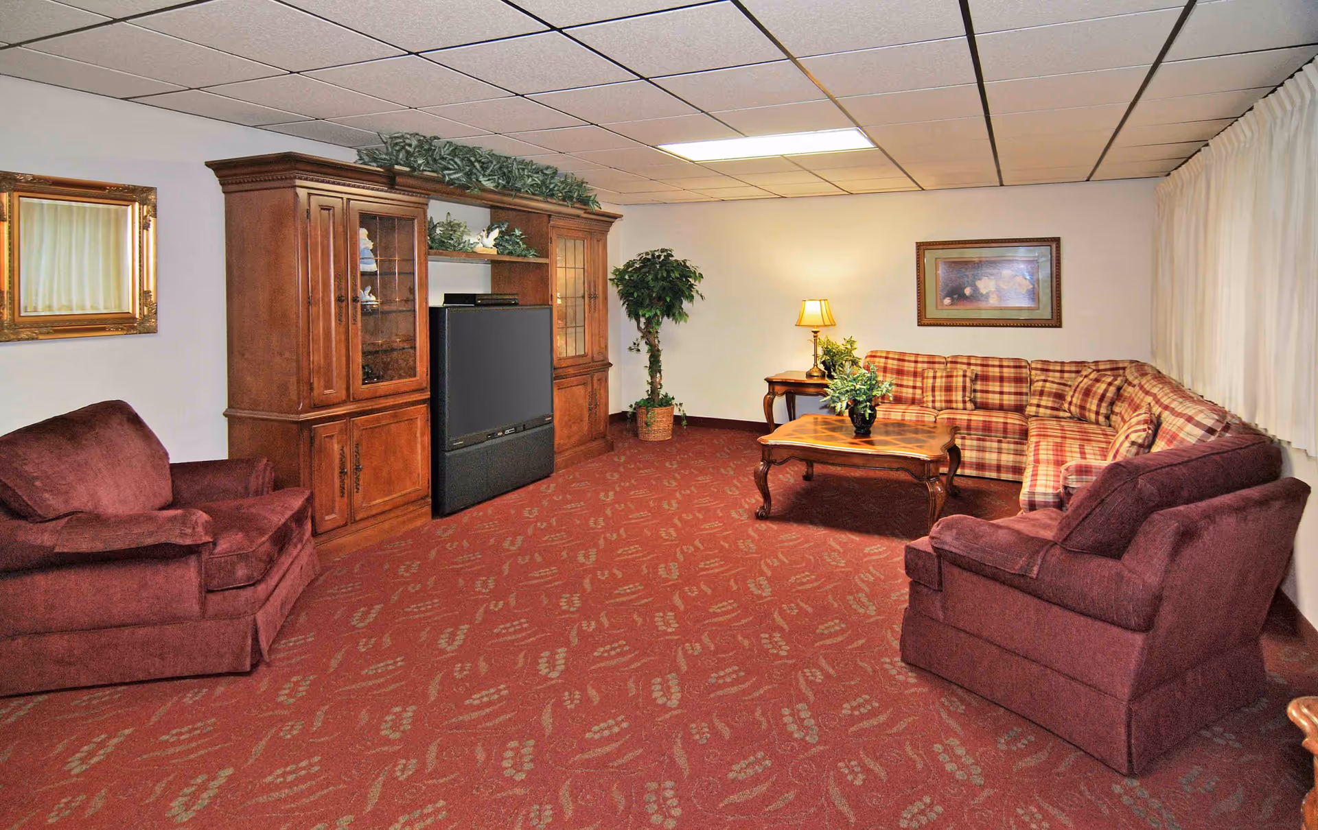 A cozy living room with a red patterned carpet, two maroon armchairs, a large wooden entertainment center with a television, a plaid sectional sofa, a wooden coffee table with a plant, a side table with a lamp, a framed painting on the wall, a large potted plant, and a window covered with white curtains.