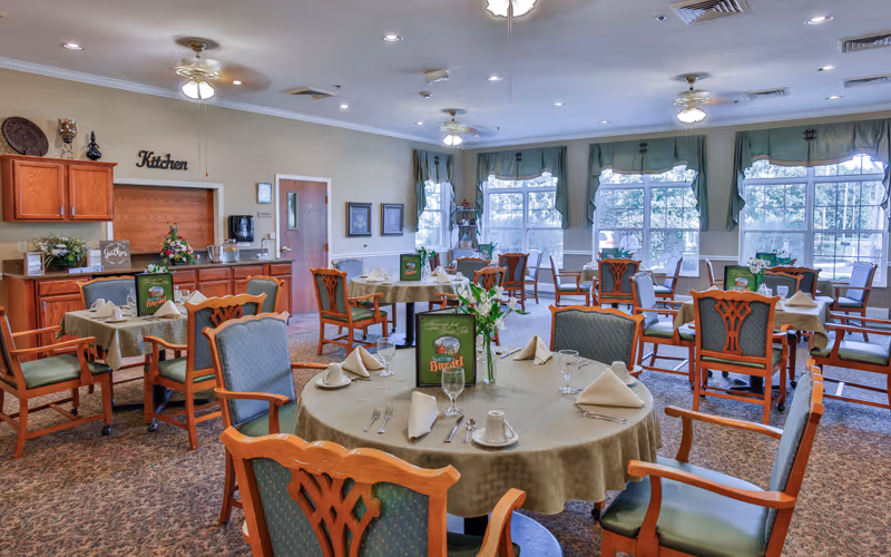 Dining room with round tables set for meals, upholstered chairs, and large windows letting in natural light.