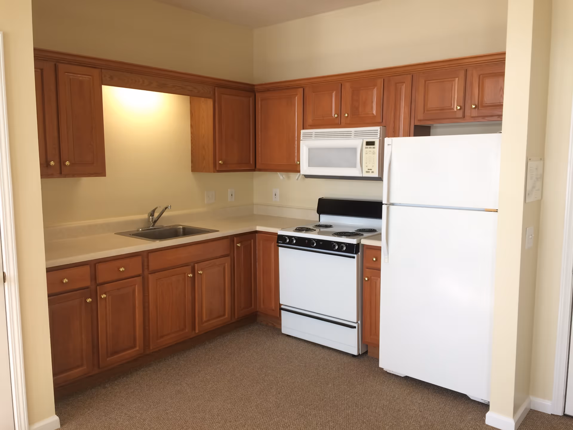 A small kitchen area with wooden cabinets, a white refrigerator, a white microwave mounted above a white electric stove, and a stainless steel sink under a cabinet with a light above it. The floor is carpeted and the walls are painted a light beige color.