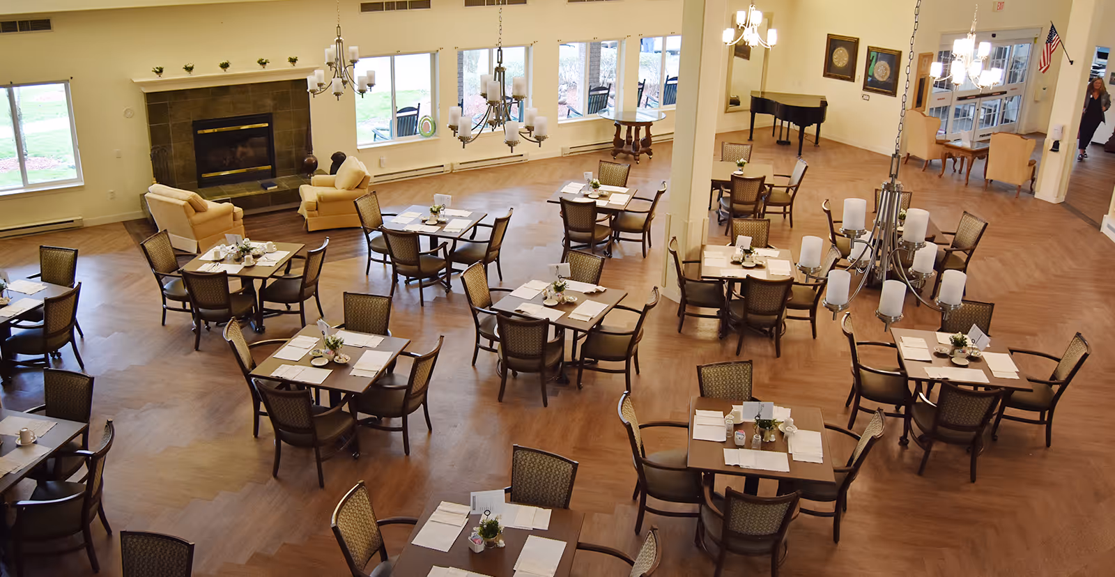 Spacious dining room with multiple square tables set with placemats, napkins, and small flower arrangements. The room features wooden flooring, large windows letting in natural light, a fireplace with a mantle decorated with small plants, chandeliers hanging from the ceiling, and a grand piano in the corner. There are also a few armchairs near the fireplace and an American flag near the entrance.
