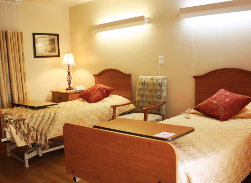 Two bedside hospital-style beds with overbed tables, patterned pillows, a nightstand with a lamp, and a chair in a warmly lit senior living room.