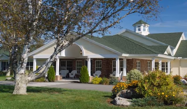 Front exterior of a single-story senior living facility with a covered porch, white columns, chairs, landscaped lawn and a green roof.
