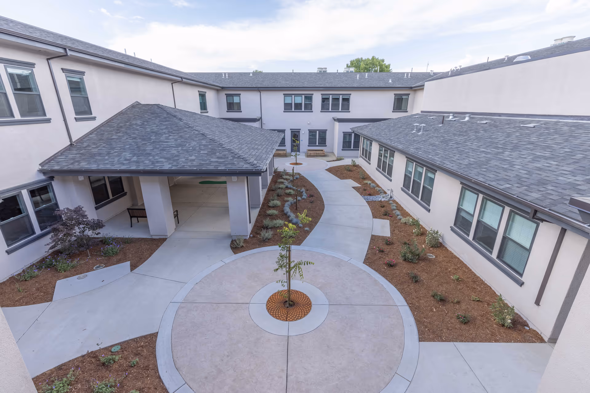 A courtyard area of a senior living facility with a circular concrete pathway surrounding a small tree in the center. The courtyard is bordered by two-story buildings with multiple windows. There are landscaped garden beds with small plants and shrubs along the pathways. A covered seating area with benches is visible on the left side.