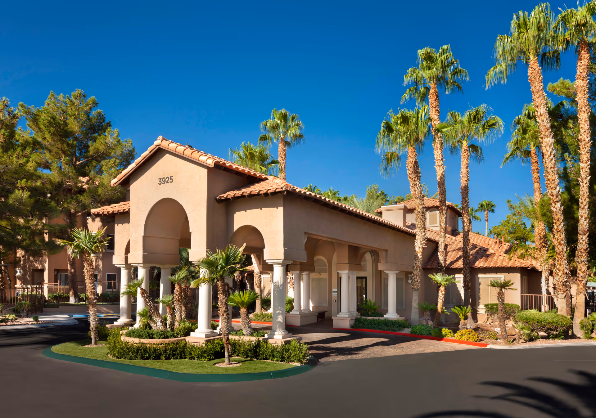 Mediterranean-style building entrance with an arched portico, columns and palm trees under a bright blue sky.