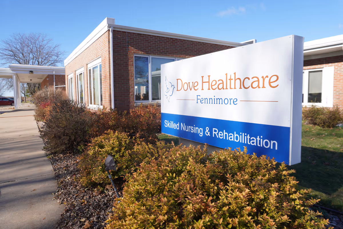 Exterior view of Dove Healthcare - Fennimore building with a large sign in front that reads 'Dove Healthcare Fennimore Skilled Nursing & Rehabilitation'. The building is made of brick and there are bushes and a sidewalk in front of it under a clear blue sky.