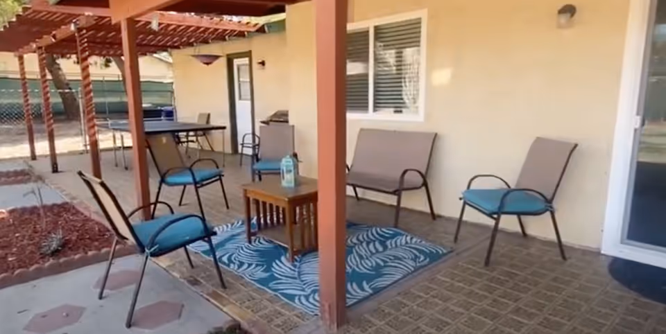 Outdoor patio area with a pergola overhead, featuring several chairs with blue cushions, a small wooden table with a bottle on it, a blue patterned rug, and a ping pong table in the background next to a beige building with windows and a door.