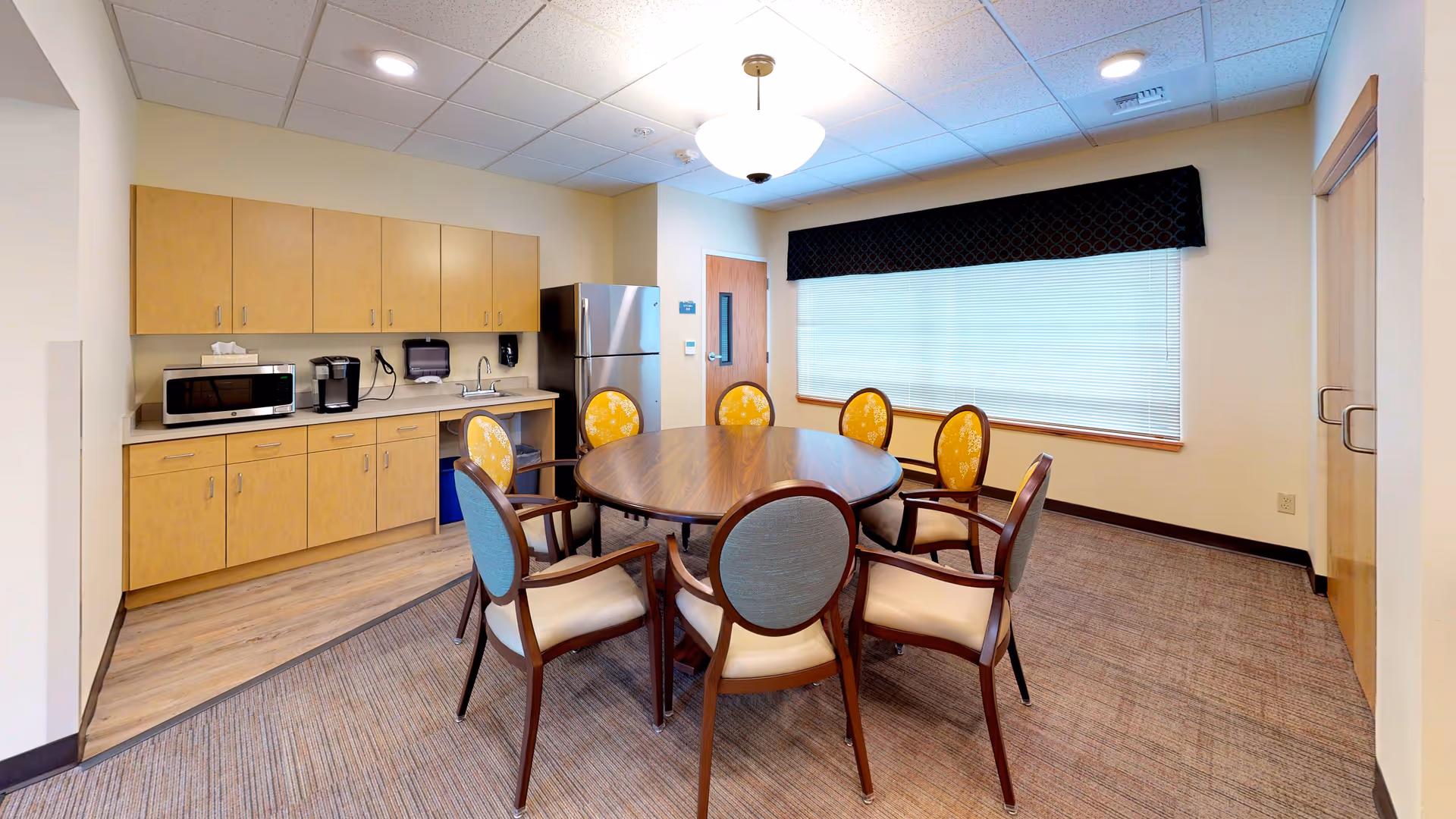 A round dining table with eight chairs sits in a bright room beside a kitchenette with cabinets, microwave, coffee maker, and refrigerator.