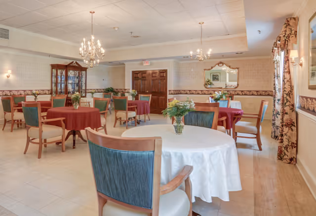 A bright dining room with round tables covered in white and red tablecloths, wooden chairs, chandeliers, floral centerpieces, and a china cabinet.