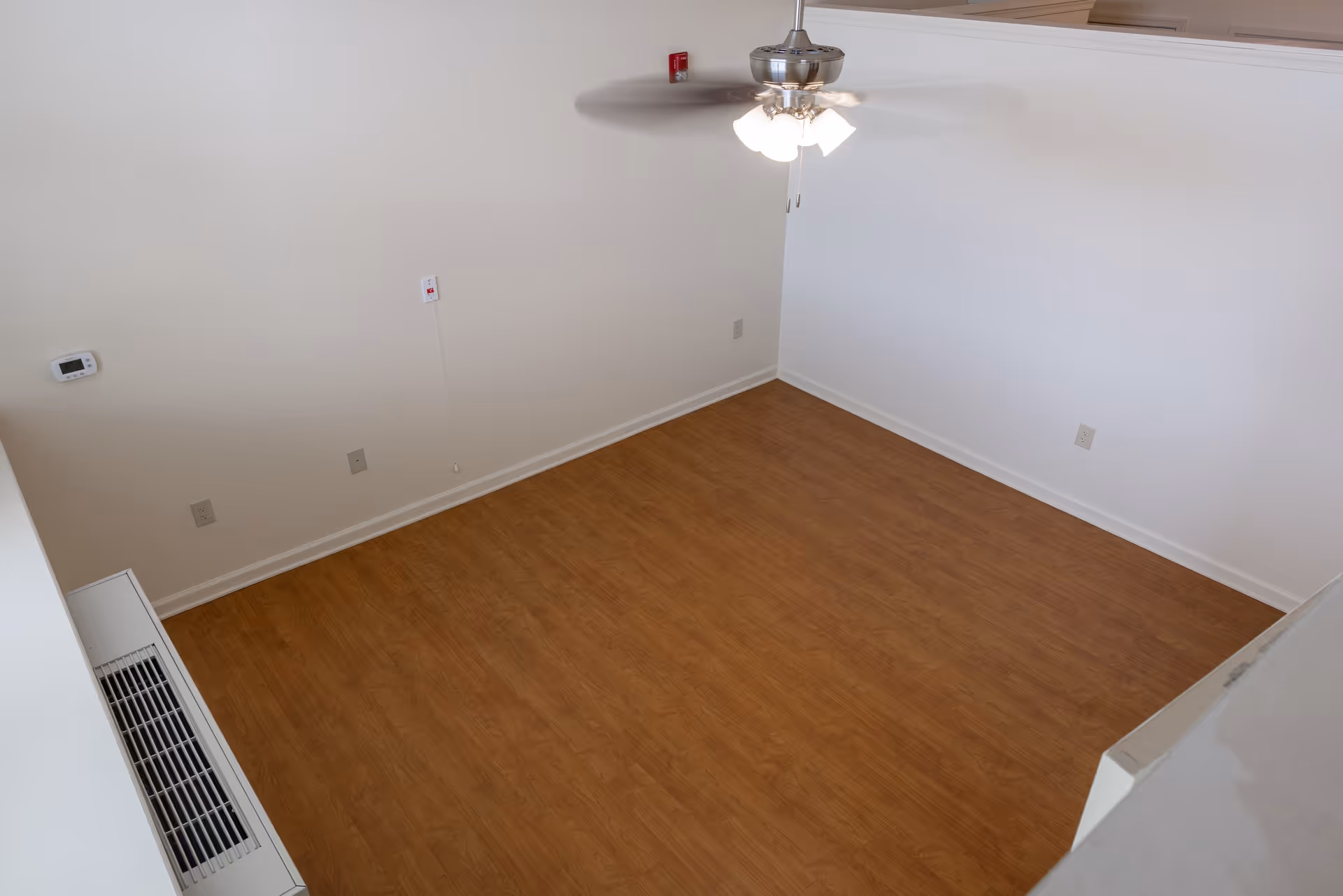 Empty room with wood flooring, white walls, a ceiling fan with lights, a thermostat on the wall, and several electrical outlets.