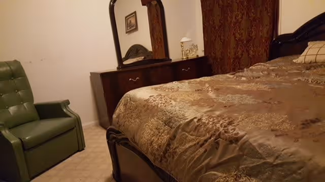 A bedroom with a large bed covered in a patterned brown and gold bedspread, a dark wooden dresser with a mirror, a small lamp on the dresser, and a green upholstered armchair in the corner.