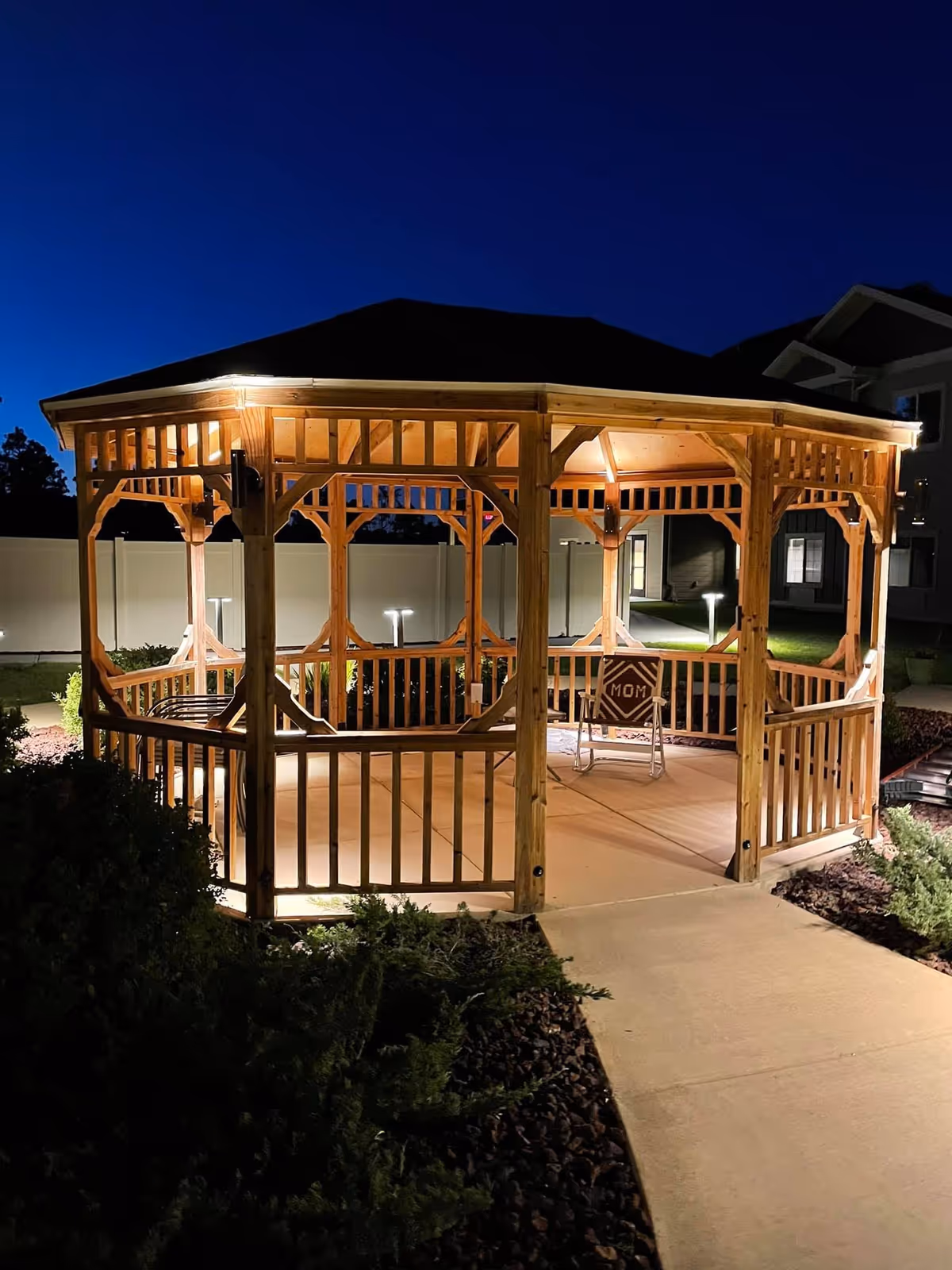 A wooden gazebo illuminated at night in a landscaped courtyard with a concrete path leading to it.