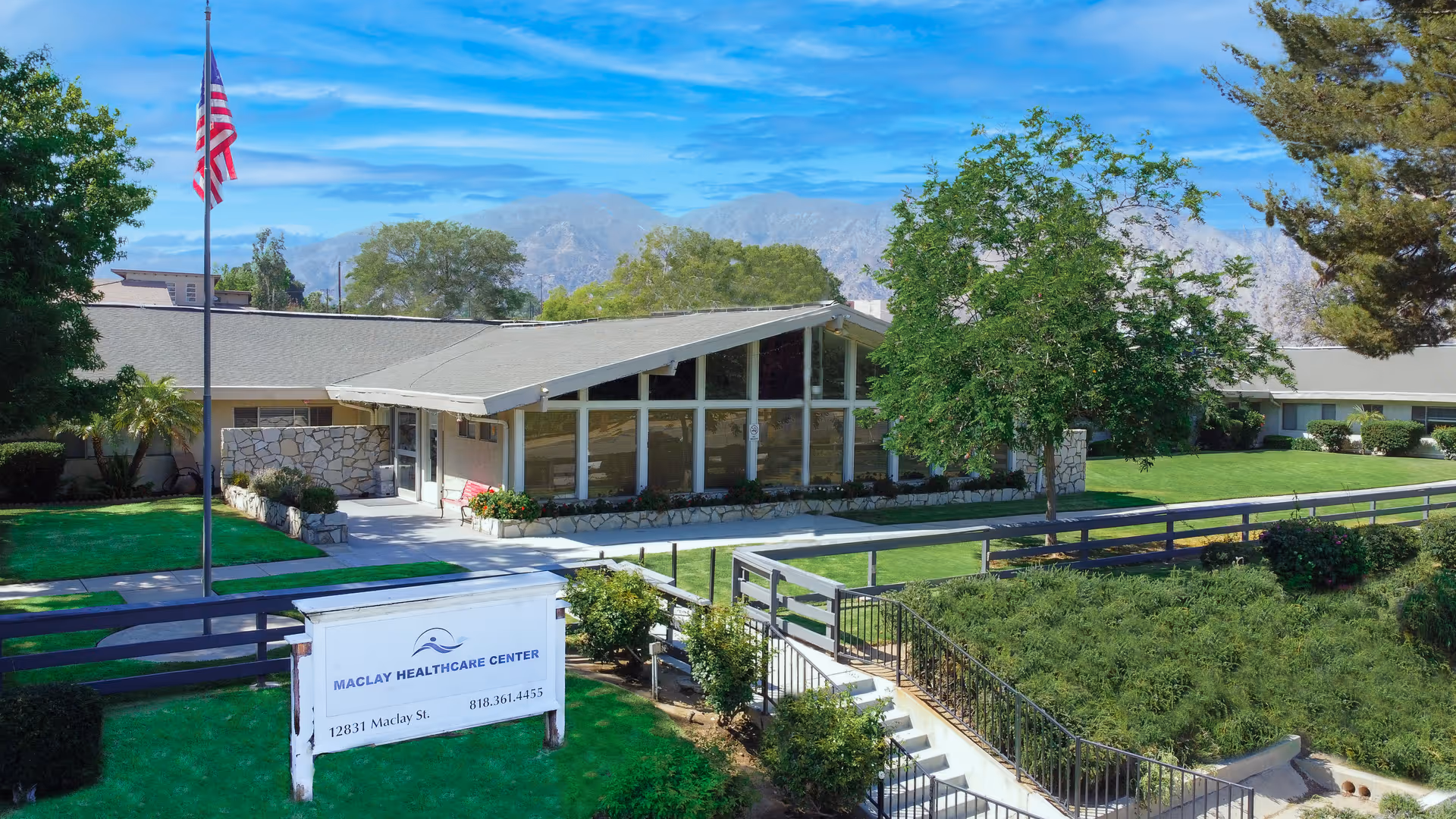 Exterior view of Maclay Healthcare Center showing a single-story building with large windows, a slanted roof, and a well-maintained lawn with trees and bushes. An American flag is flying on a flagpole near the entrance, and a white sign with the facility's name and contact information is visible in front.