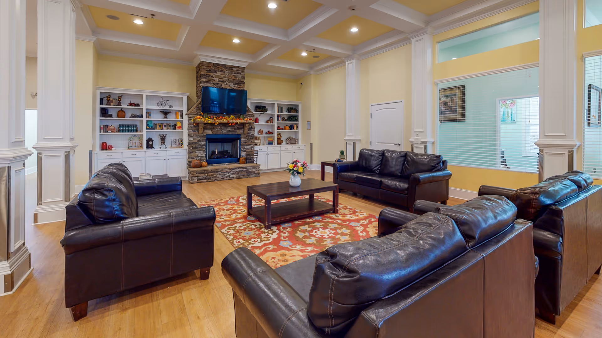 Spacious communal living room with multiple leather sofas arranged around a coffee table on a patterned rug, a stone fireplace with a TV, and built-in white shelving.