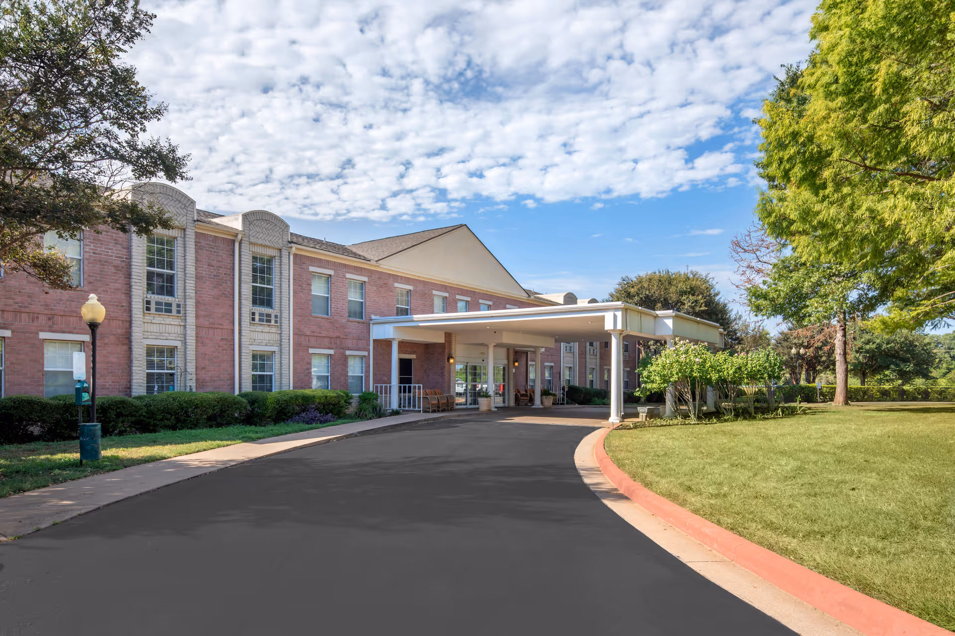 Front entrance of a two-story brick senior living building with a covered drop-off porte-cochère, driveway, and landscaped lawn under a partly cloudy sky.