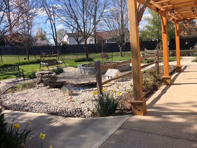 Outdoor courtyard with a pergola-covered walkway, pebble garden and small stone water feature, benches, and trees.