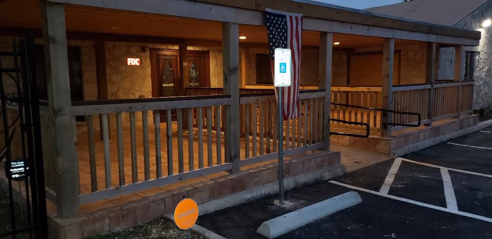 Evening view of a covered wooden porch with railings and a ramp leading to double wooden doors with glass panels. An American flag hangs from a pole near a handicapped parking sign in front of the porch. The area is lit with warm lighting and there is a parking lot with marked spaces adjacent to the porch.